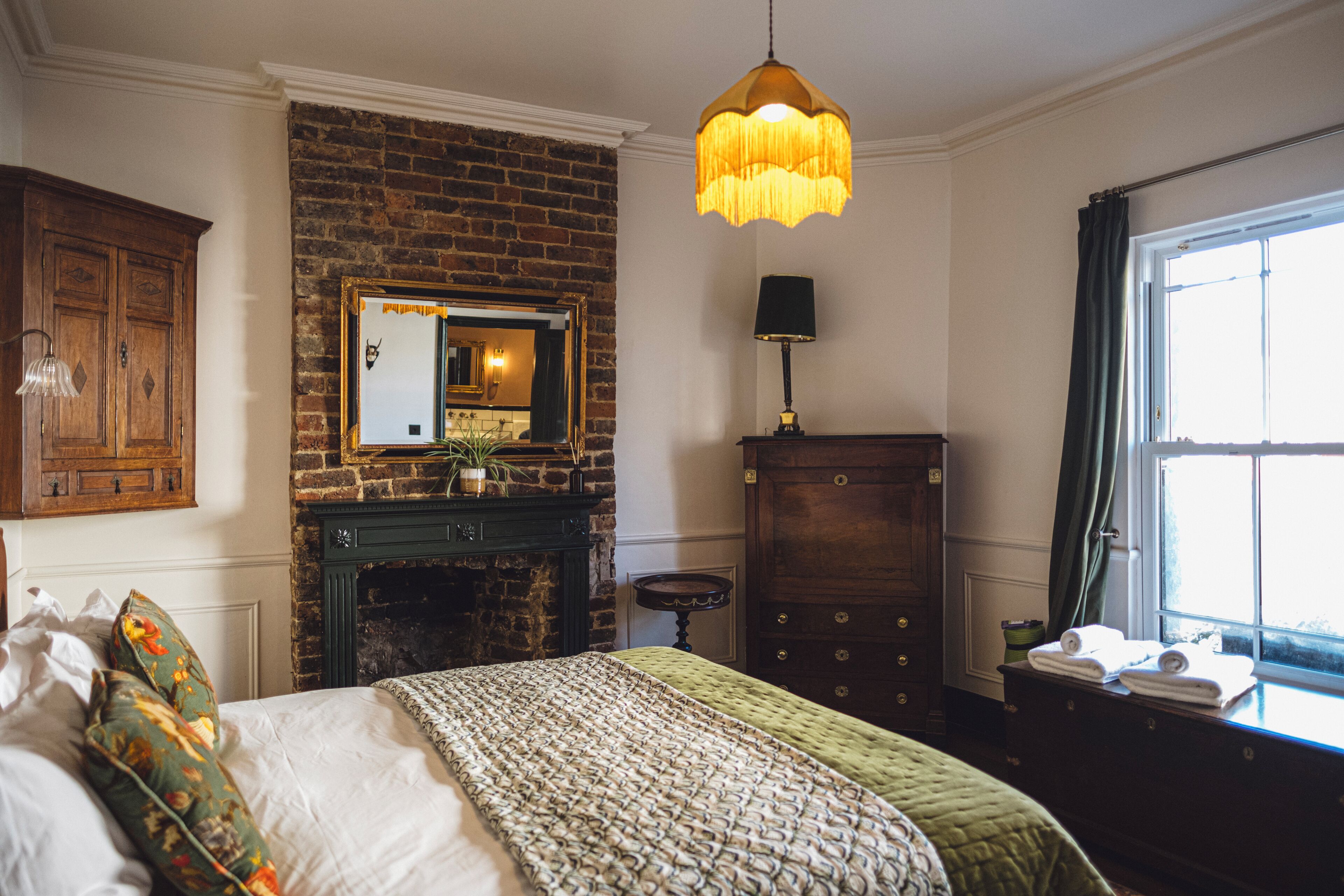 Un dormitorio con chimenea de ladrillo, cama con almohadas florales, lámpara colgante y cómoda con toallas.