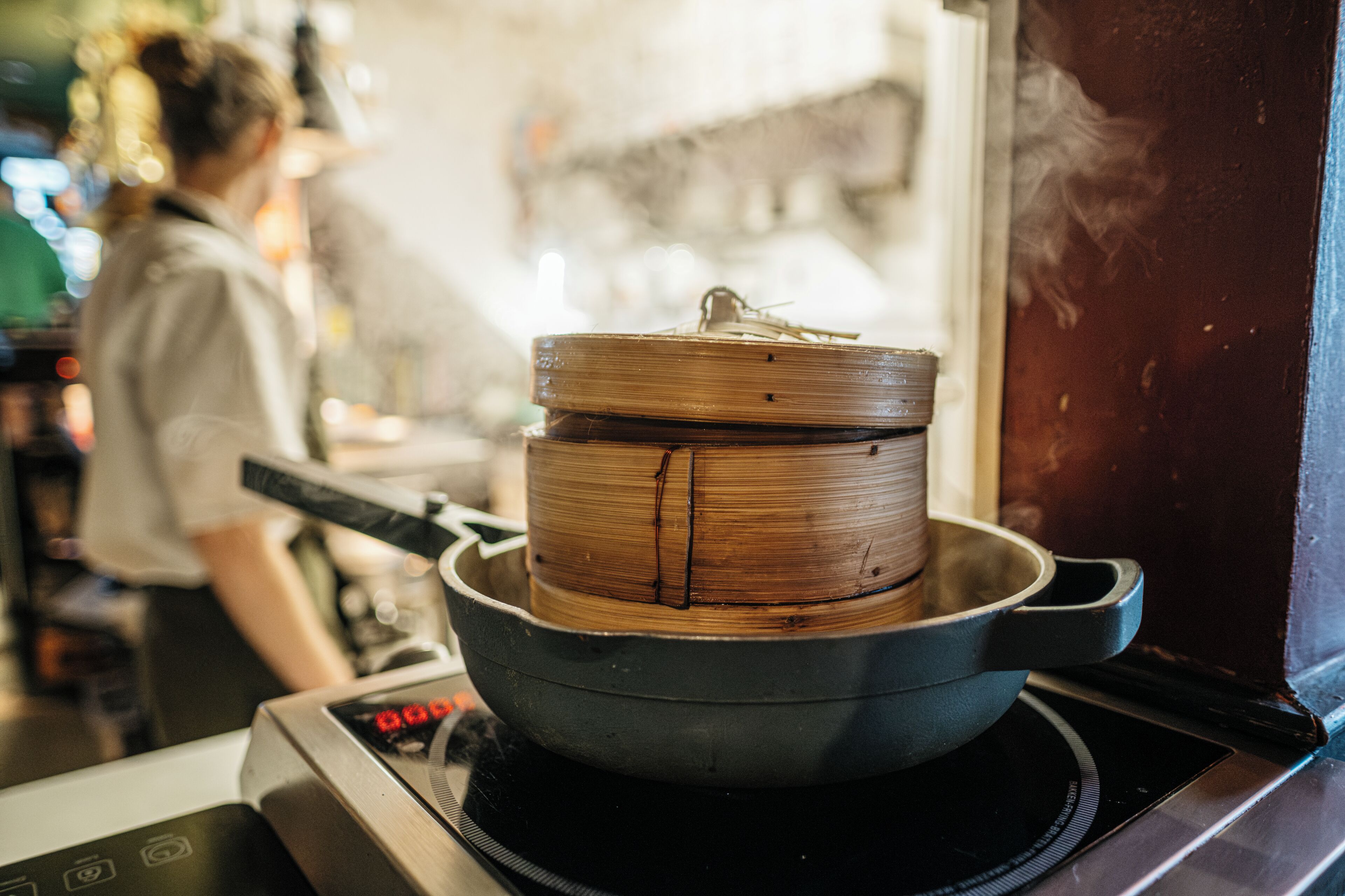 Une casserole sur une cuisinière, avec un couvercle, de la vapeur s'en échappant, et une personne à l'arrière-plan.