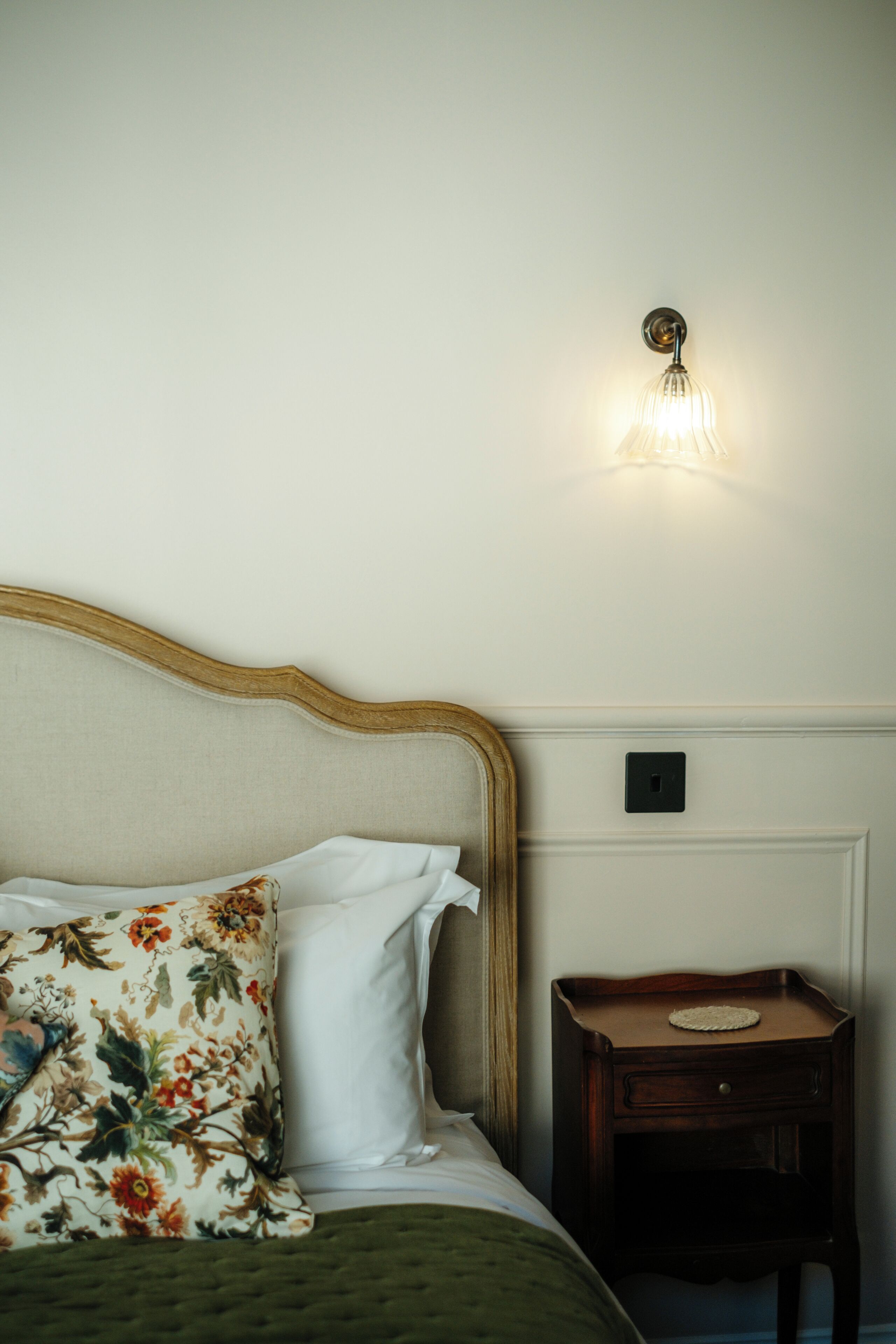 Un lit bien fait, agrémenté d’oreillers à motifs floraux, une table de chevet avec une lampe et un interrupteur mural.