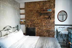 A bedroom with a brick wall, a bed with white linens, a small shelf with books, and a round mirror. - The Doghouse Inn (Hastings)