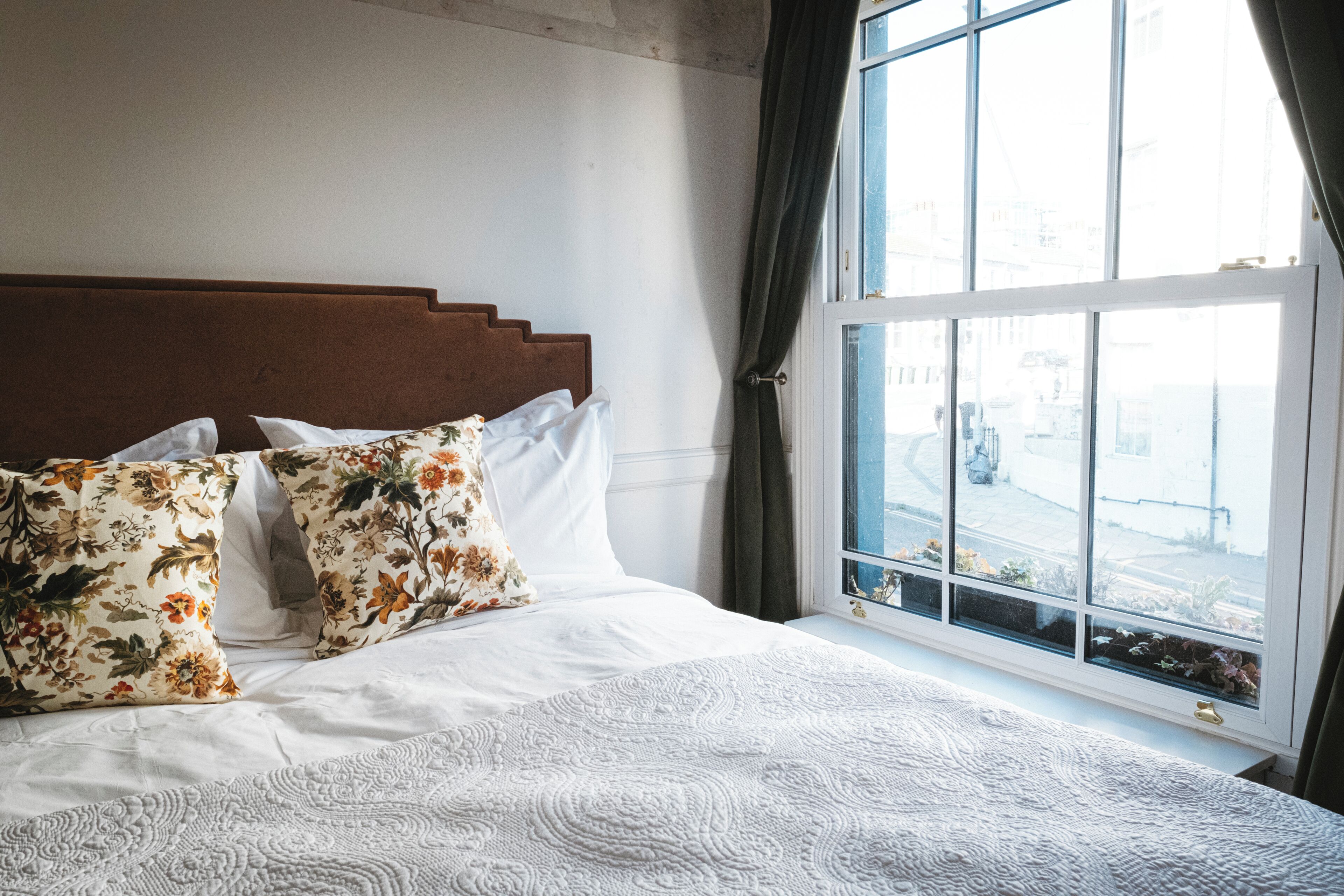 A bed with floral pillows, a white quilt, and a view of a city through a window.