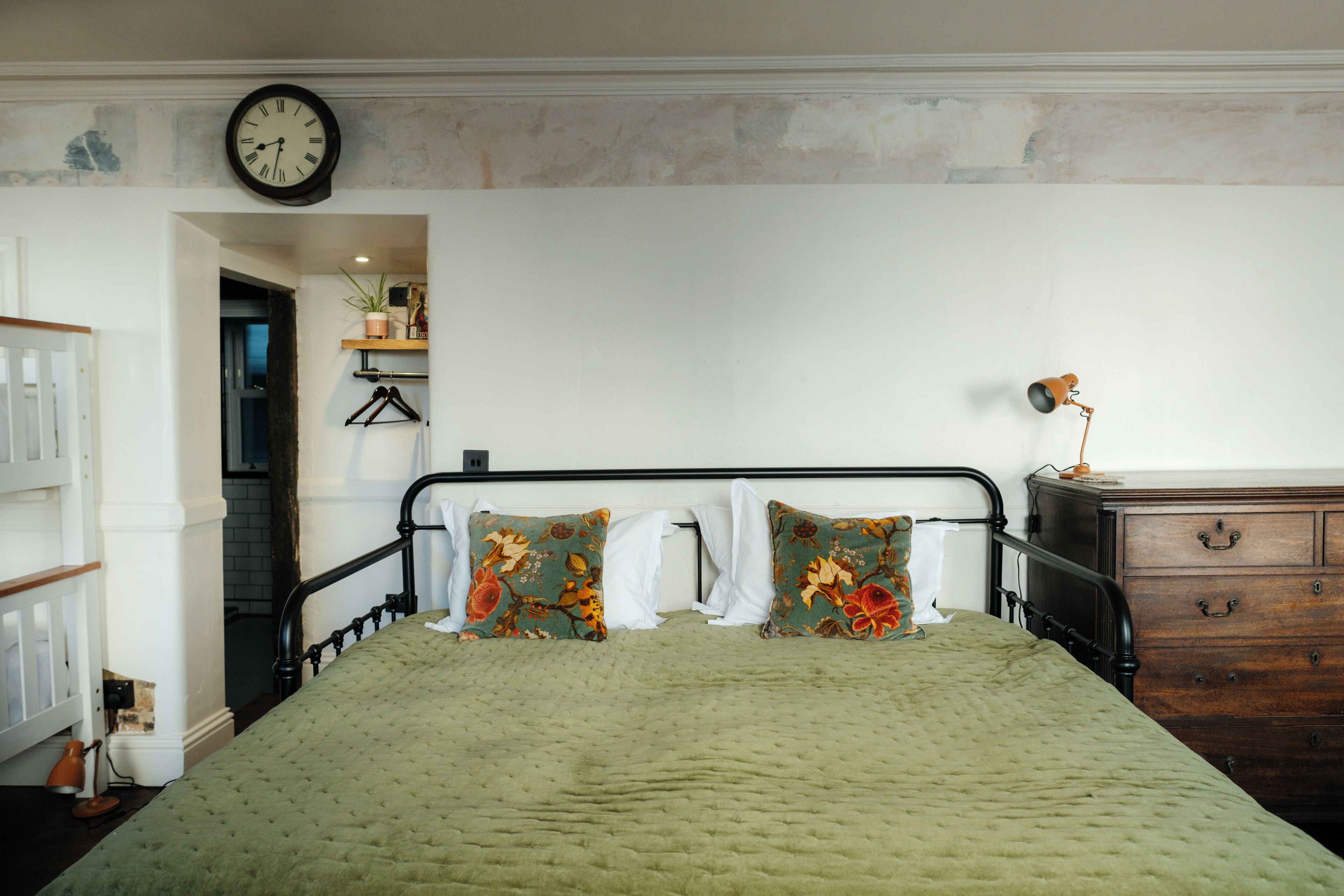A bedroom with a green quilted bedspread, two decorative pillows, a wooden dresser, and a wall clock.