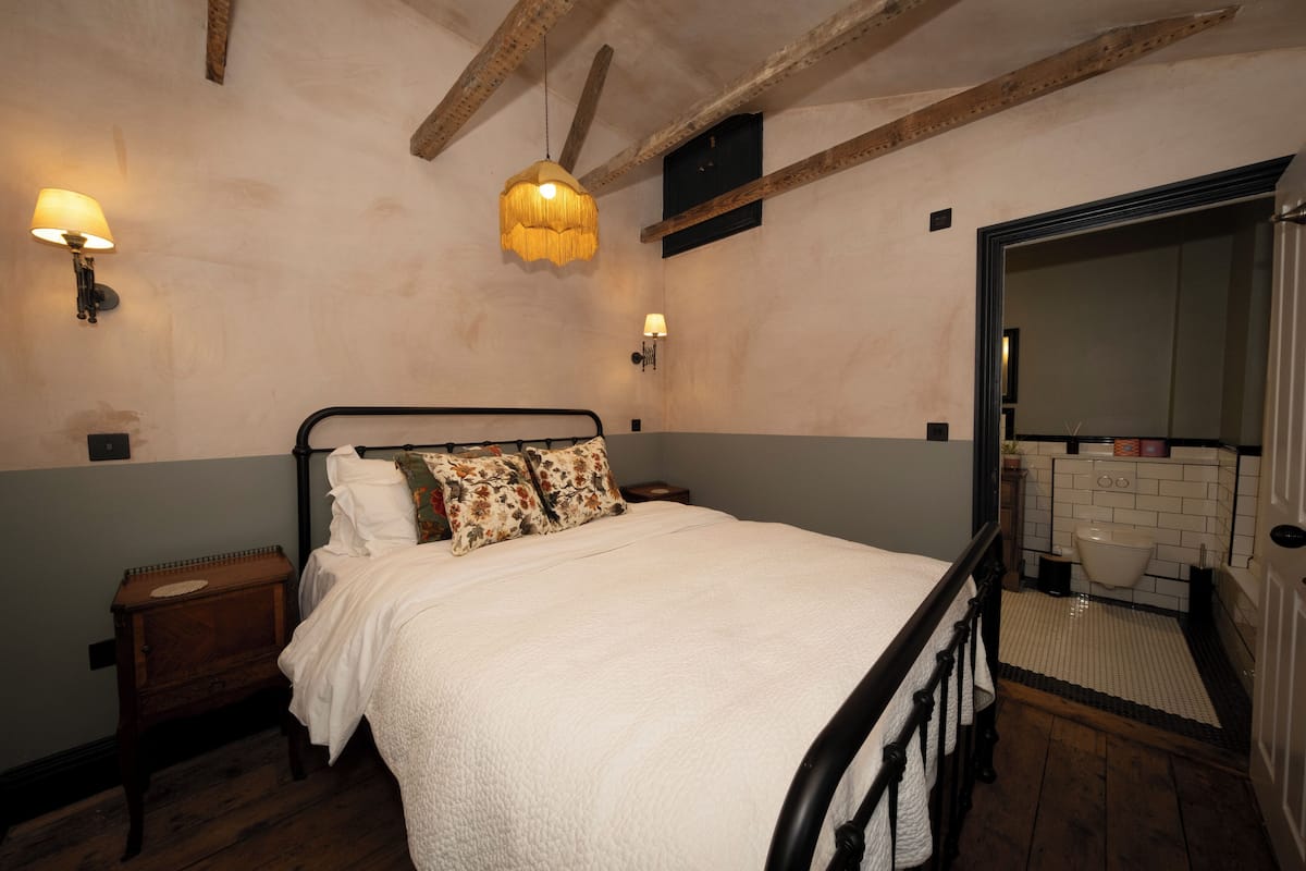 A bedroom with a bed, nightstand, wall-mounted lamps, and a bathroom visible through an open door.