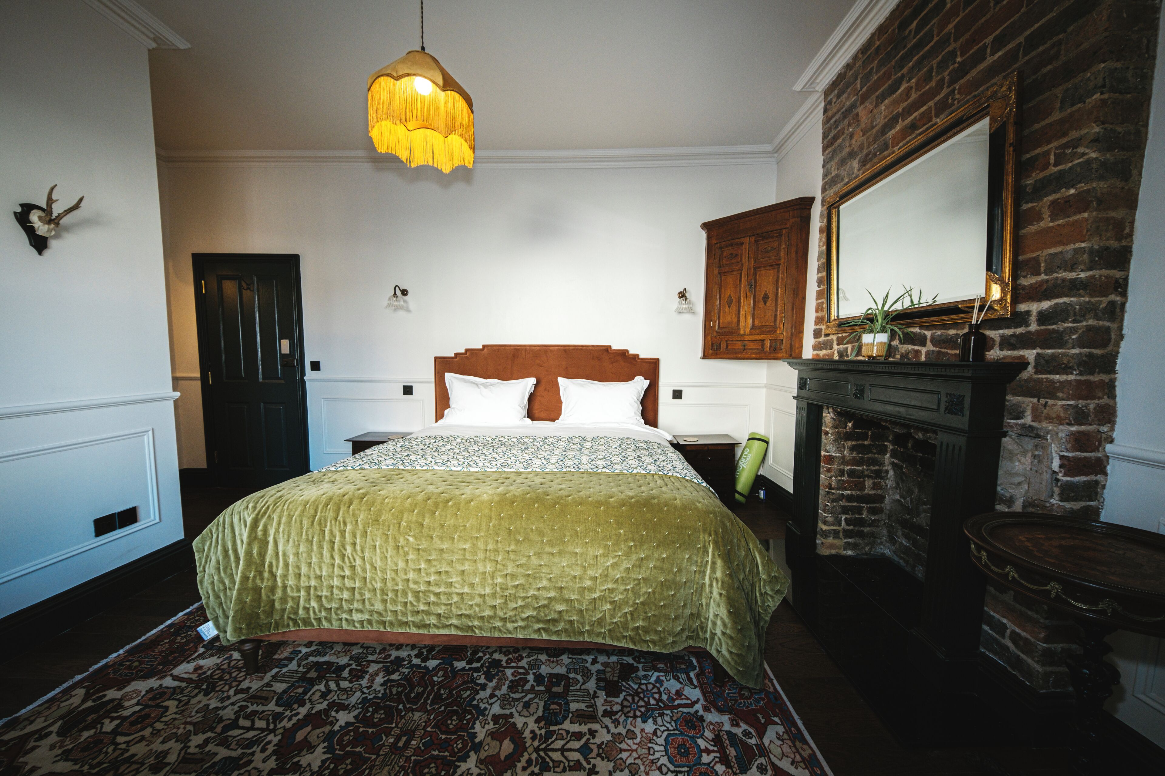 A bedroom with a large bed, a wooden headboard, a hanging light fixture, a brick fireplace, and a mirror above it.