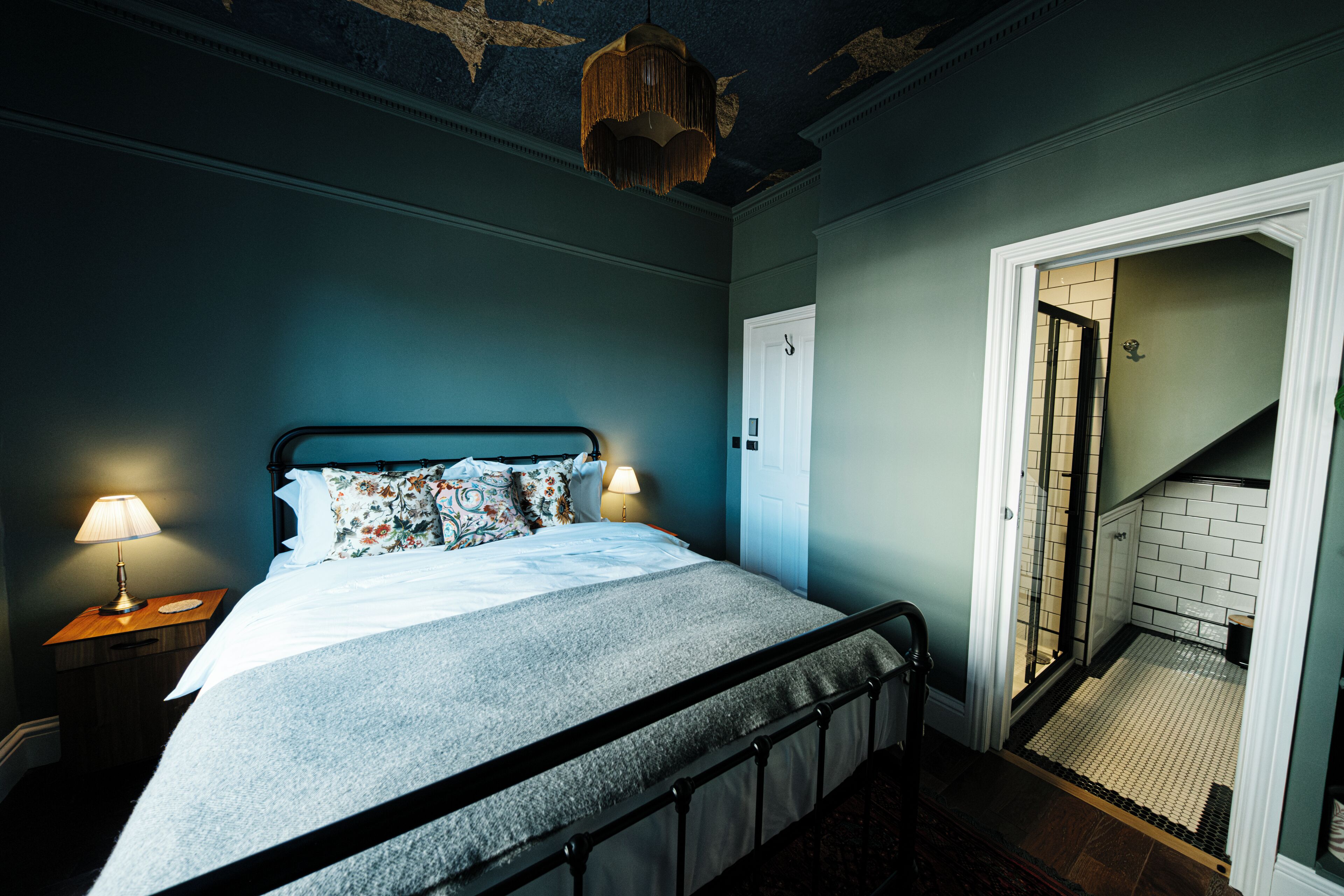 A bedroom with a bed, nightstands, a hanging light fixture, and a door leading to a bathroom.