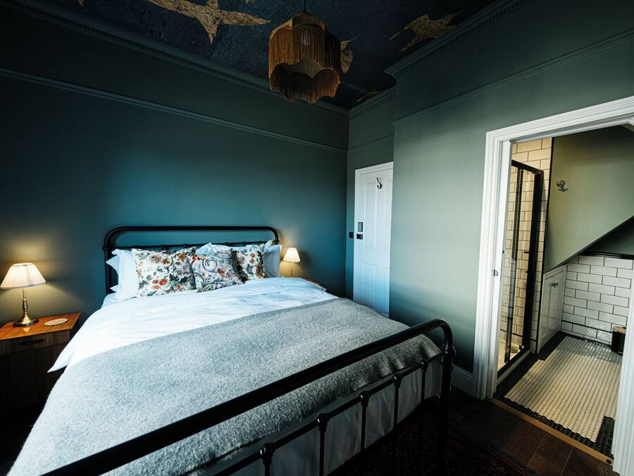 A bedroom with a bed, nightstands, a hanging light fixture, and a door leading to a bathroom.