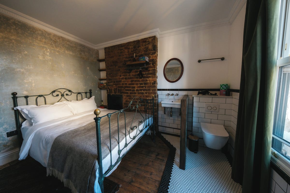 A bedroom with a brick wall, a bed with white linens, a wooden floor, a window with curtains, and a bathroom with a white sink and toilet.