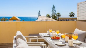 Una tavola imbandita per la colazione, con croissant, frutta e succo d'arancia, con vista su una spiaggia, palme e un cielo azzurro terso.