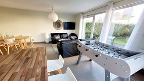 A modern living room with a foosball table, a dining area with a wooden table and chairs, a sofa, and a large window with curtains. - Modern villa 3 bedrooms with garden, WiFi and parking, close to beach (Wimereux)