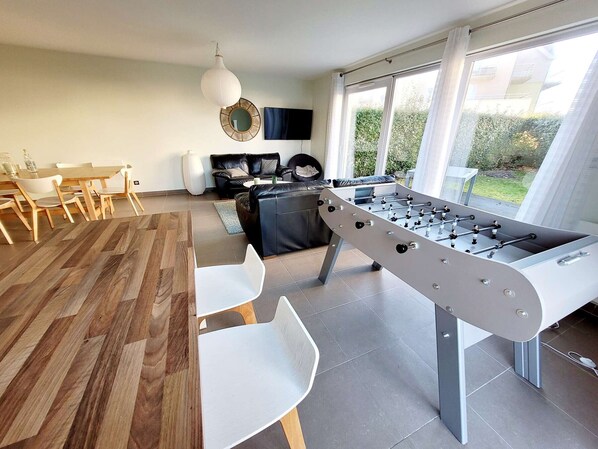 A modern living room with a foosball table, a dining area with a wooden table and chairs, a sofa, and a large window with curtains. - Modern villa 3 bedrooms with garden, WiFi and parking, close to beach (Wimereux)