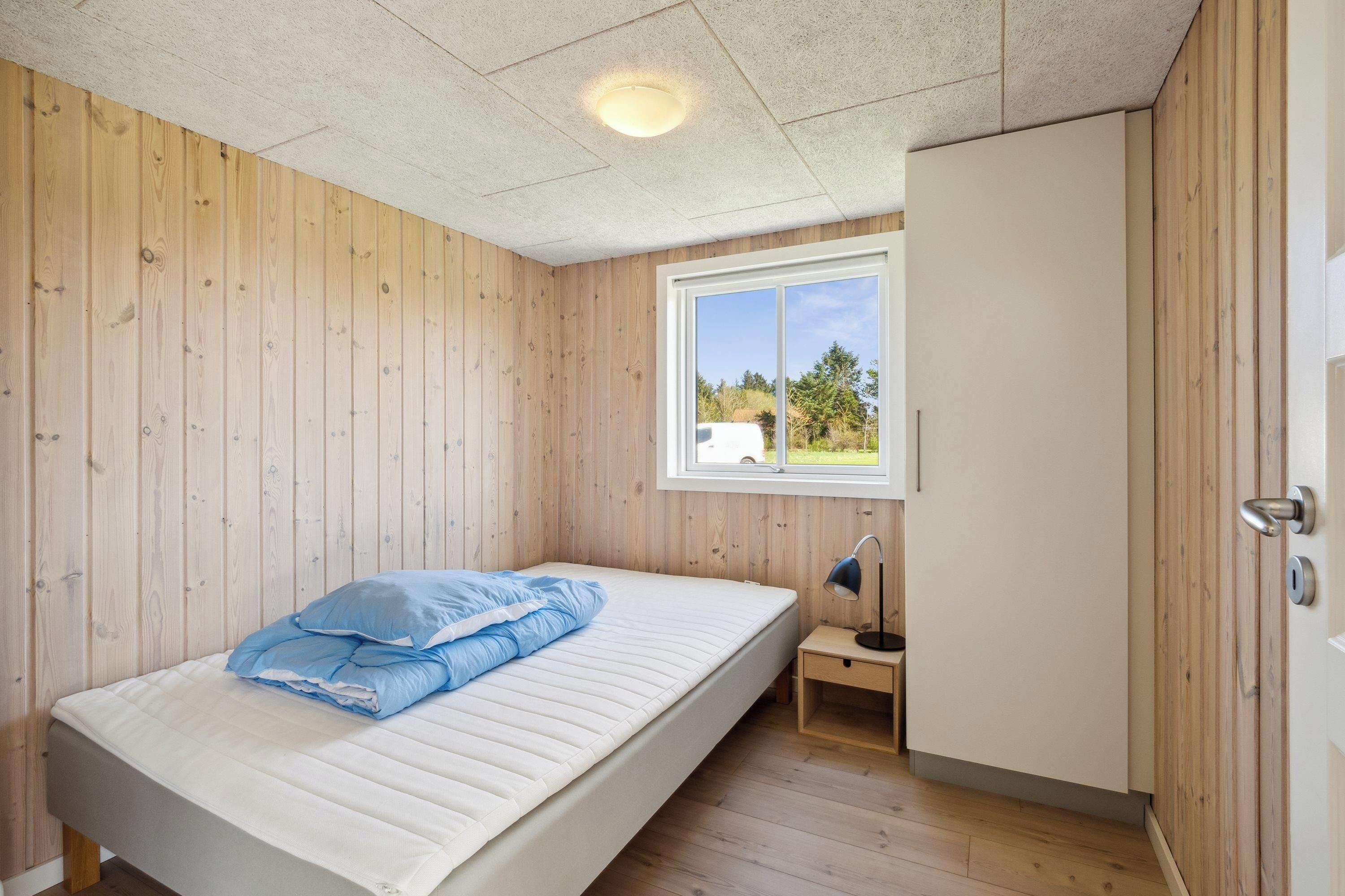 Quarto individual com paredes de madeira, janela com vista para o verde, mesa de cabeceira com abajur e guarda-roupa.