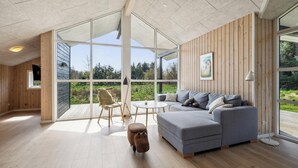 A modern living room with a grey sofa, wooden flooring, large windows, and a wooden wall panel. - Modern holiday home from 2017, located at Lild Strand. (Frøstrup)