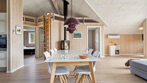 A spacious room with a large dining table, white chairs, and a modern pendant light. A fireplace is visible in the background, and there is a sofa in the corner. - Modern holiday home from 2017, located at Lild Strand. (Frøstrup)