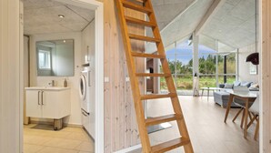A modern interior with a wooden ladder leaning against a wall, a bathroom with a washing machine, and a seating area with a view of the outdoors. - Modern holiday home from 2017, located at Lild Strand. (Frøstrup)