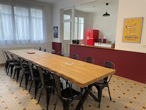 Un comedor con una larga mesa de madera y sillas negras, un refrigerador rojo, y una ventana con cortinas translúcidas.