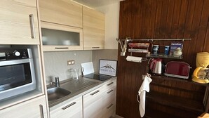 Kompakti keittiö, jossa on sisäänrakennettu uuni, pesuallas ja pieni työtaso. Siellä on myös puinen kaappi lasiovella, hylly keittiövälineille ja kahvinkeitin.