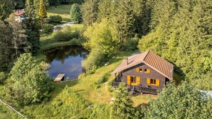 Una cabaña de techo inclinado, rodeada de un bosque frondoso y un pequeño estanque.