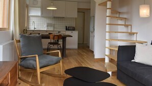 A modern living room with a wooden rocking chair, a dark sofa, and a black ottoman. A kitchen with white cabinets and a hanging light is visible in the background. - 4-room apartment for 6 guests, parking and balcony in Méribel (Méribel)