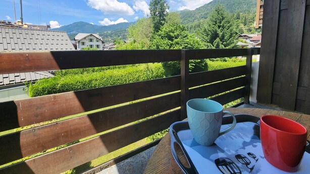 En balkon med trægelænder, en bakke med to krus og udsigt over et bjergegnskab med huse og en mobilkran.