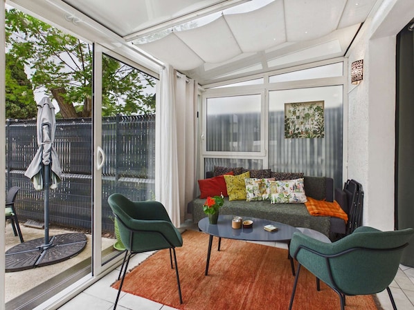 A sunroom with a glass door, a round table, two chairs, a sofa with colorful cushions, and a potted plant.