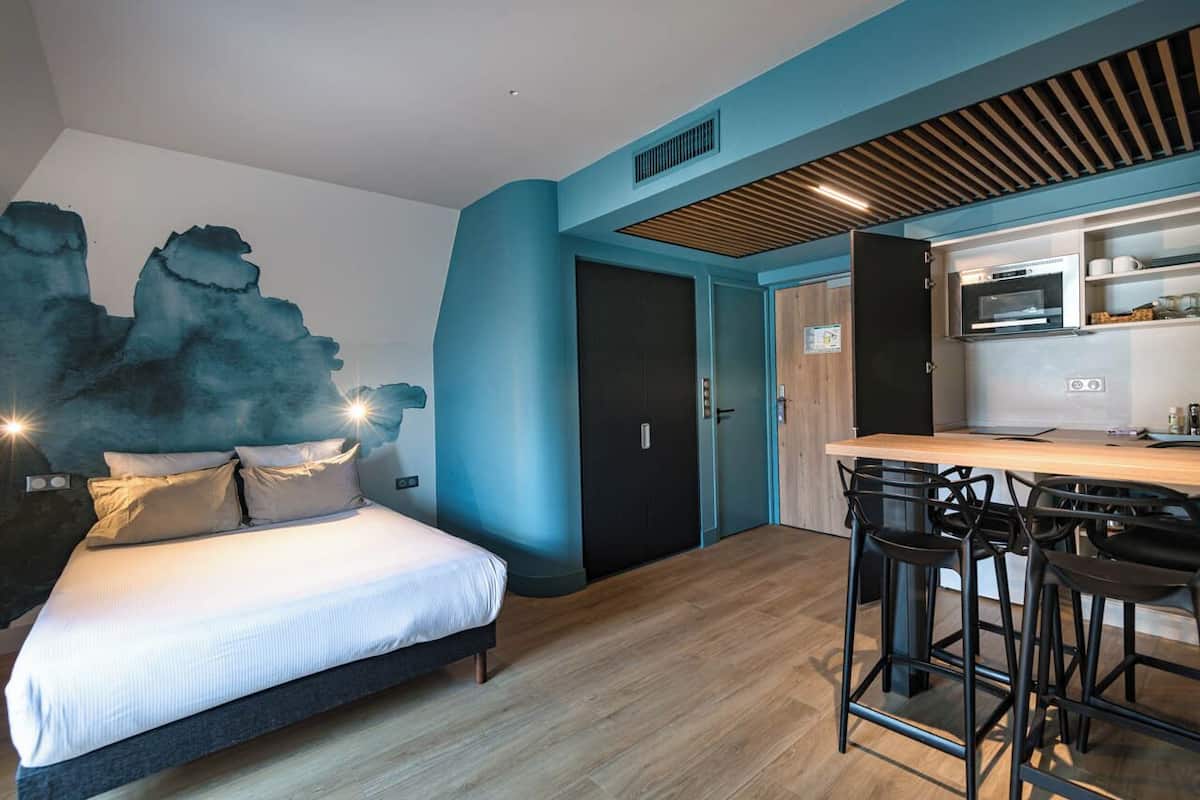 Un dormitorio moderno con cama, una zona de cocina con mostrador y taburetes, y una puerta que da a otra habitación.