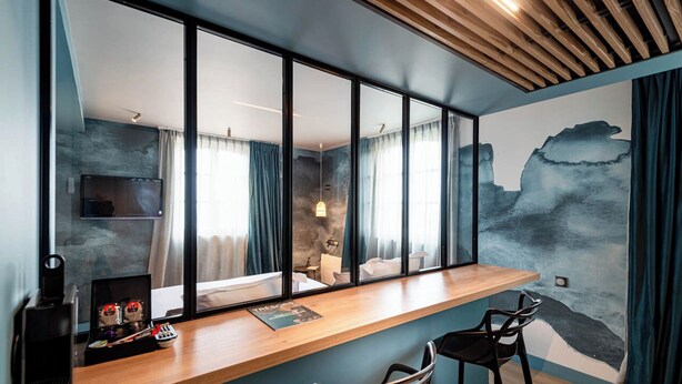 A modern room with a large window, a wooden desk, two bar stools, and a painting on the wall.