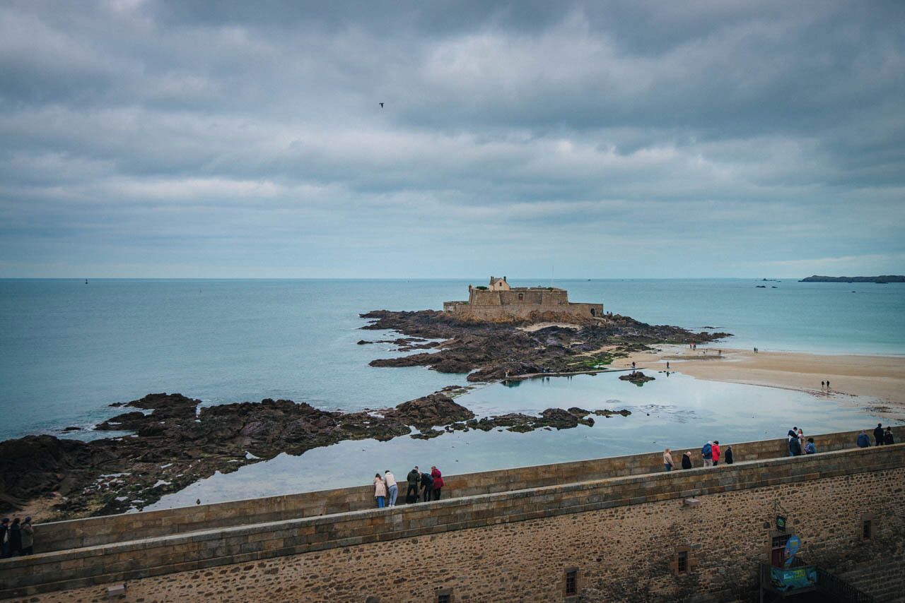 A coastal view with a historic stone structure on rocky outcrops, a sandy beach, and a stone wall with people.