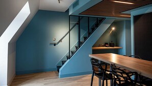 A modern interior with a blue wall, a wooden staircase, a dining table with chairs, and a small kitchen area.