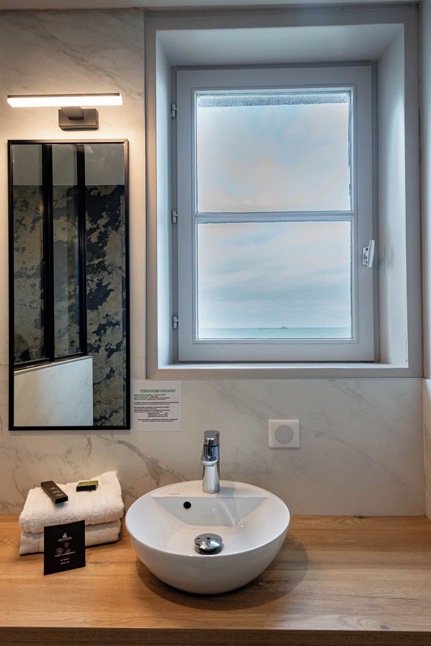 A bathroom with a round white sink, a mirror with a camouflage pattern, and a window with a view of the sea.