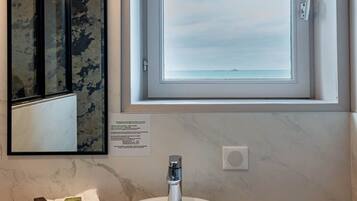 A bathroom with a round white sink, a mirror with a camouflage pattern, and a window with a view of the sea.