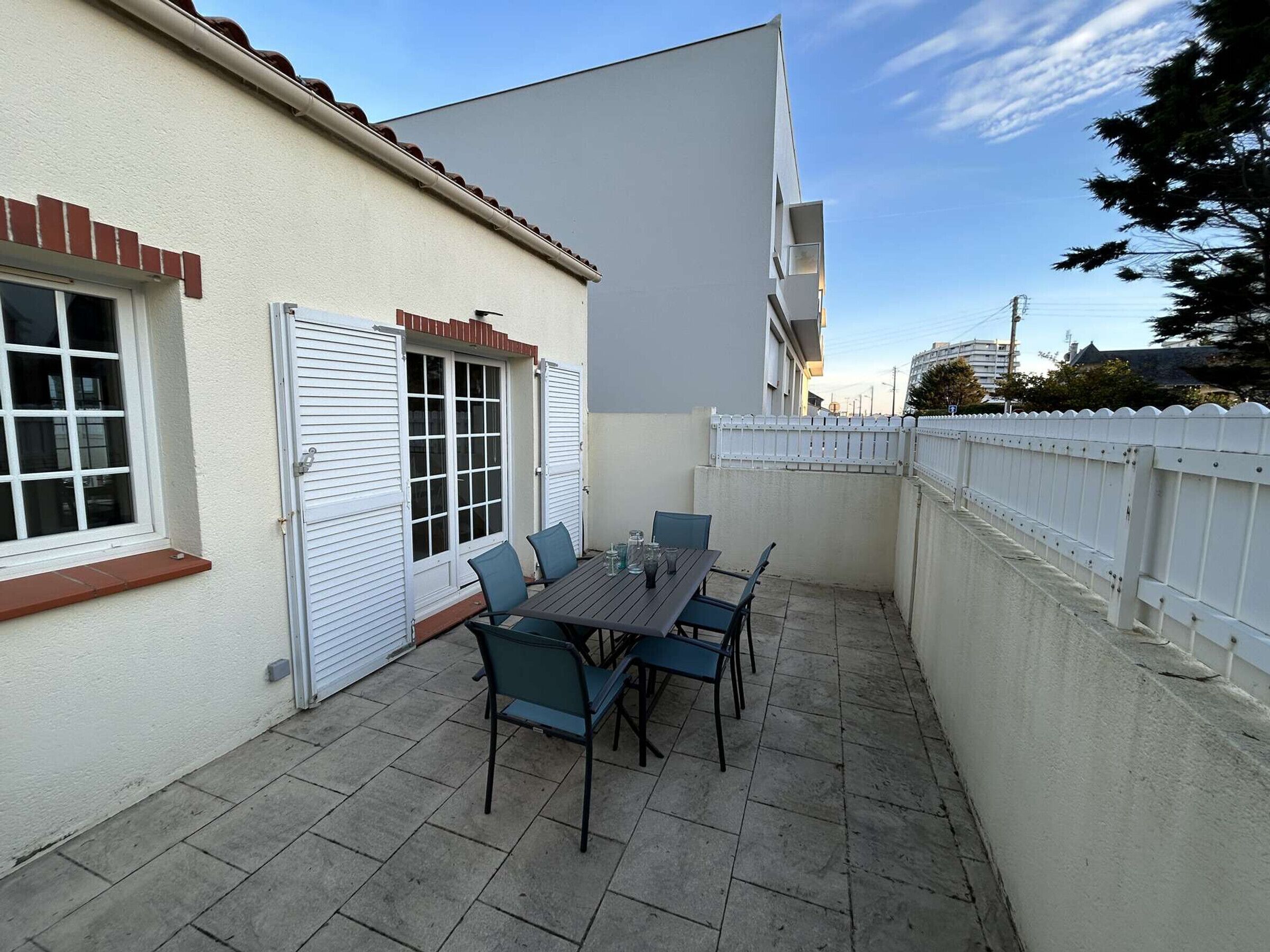 Un patio con una mesa y sillas, cerca de una valla blanca y un edificio con ventana y contraventanas.