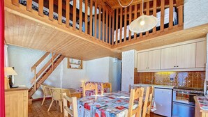 A cozy room with a wooden staircase, a dining table with chairs, a kitchen area, and a bed with a patterned bedspread. - Cozy Duplex in La Clusaz - 5 pers, pets allowed (La Clusaz)