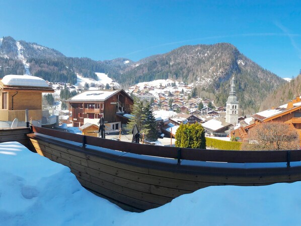A wooden boat on a snowy surface, overlooking a mountainous village with buildings and a church.
