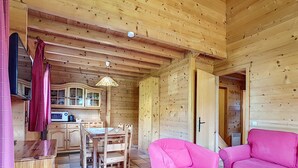 A wooden cabin interior with a dining table, chairs, a pink sofa, and a kitchen area.