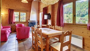 A wooden cabin with a dining area, two pink chairs, a fireplace, and large windows overlooking a forest.
