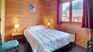 A wooden cabin bedroom with a bed, two bedside lamps, a window with curtains, and a painting on the wall.