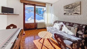 A room with a wooden floor, a sofa with a winter-themed cover, a television on the wall, and a window with a view of snow-covered hills. - Cozy studio in La Clusaz, 4 pers, pets allowed (La Clusaz)