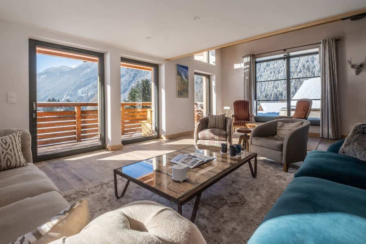 Een ruime woonkamer met een grote glazen salontafel, comfortabele zithoek en uitzicht op besneeuwde bergen door grote ramen.