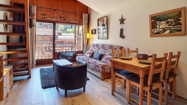 Una sala acogedora con techo de madera, un sofá estampado, una mesa de comedor con sillas y vistas a un espacio exterior a través de un ventanal.