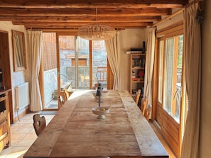 A rustic wooden dining table with a bowl of fruit, wooden chairs, and a hanging light fixture in a room with stone walls and large windows.