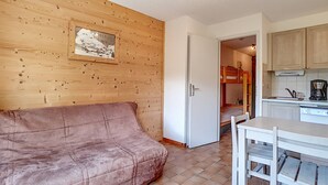 A compact living space with a wooden wall, a pink sofa, a dining table with chairs, and a kitchen area with cabinets and a coffee maker.
