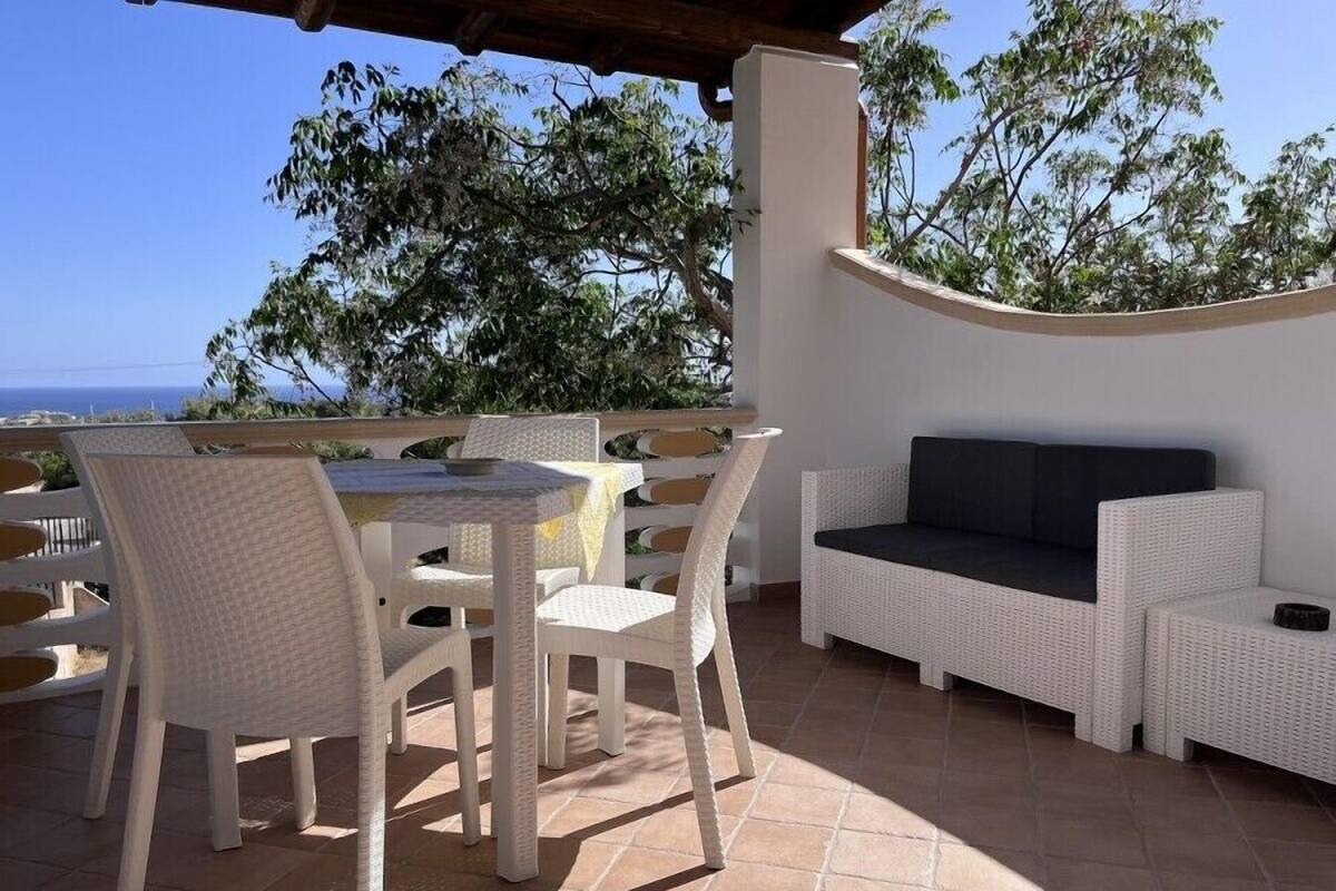 Eine Terrasse im Freien mit einem runden Tisch und Stühlen, einem weißen Korbsessel und Blick aufs Meer.