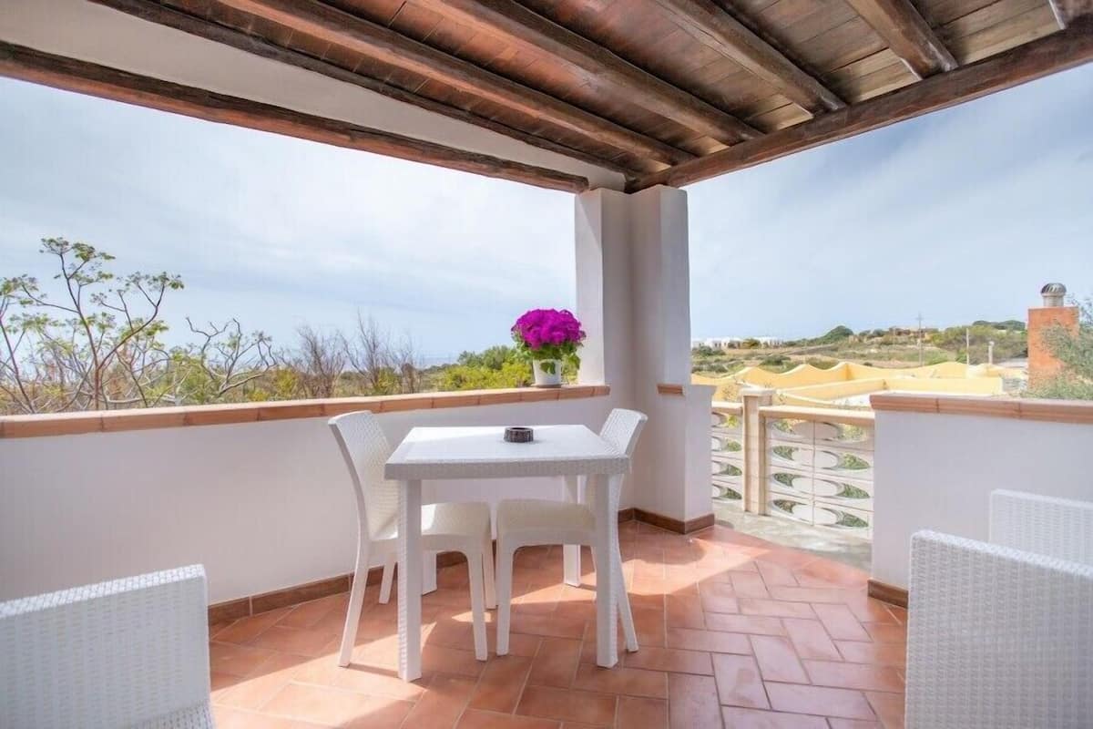 Eine überdachte Terrasse mit einem weißen Tisch und Stühlen, ein Blumentopf mit violetten Blüten und Blick auf die Landschaft.