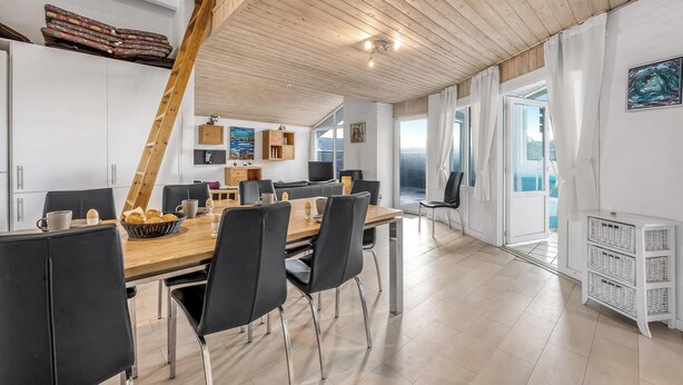 A spacious dining area with a wooden table and chairs, a kitchen in the background, and large windows offering views. - Lovely holiday home with pool, spa and sauna, only 200 meters from the North. (Harboøre)