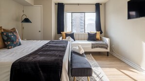 A modern bedroom with a large bed, a sofa bed, a window with curtains, and a flat-screen TV mounted on the wall.