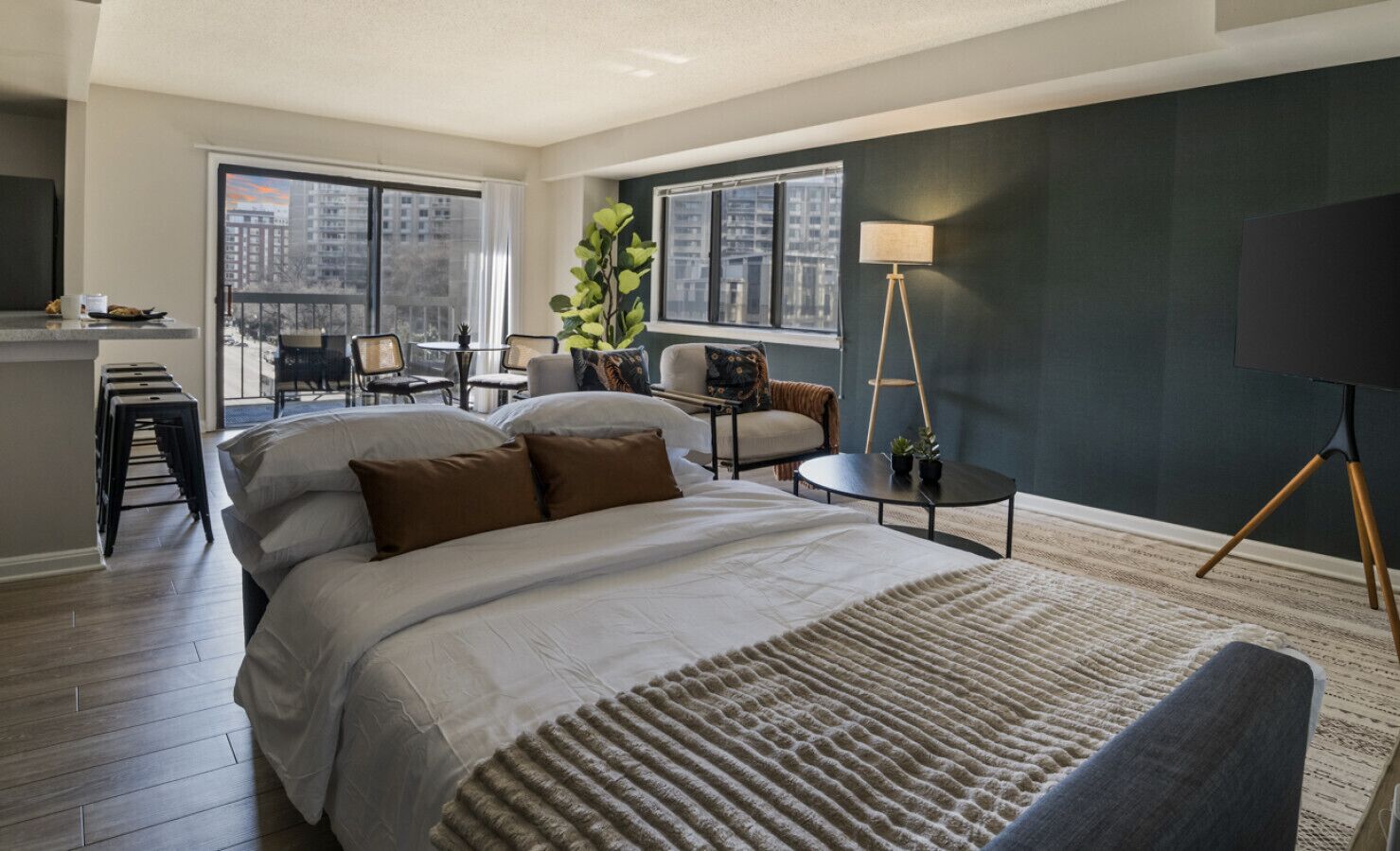 Habitación de hotel con una cama grande, un escritorio, una silla, una lámpara y vistas a la ciudad a través de amplios ventanales.