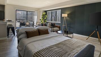 Habitación de hotel con una cama grande, un escritorio, una silla, una lámpara y vistas a la ciudad a través de amplios ventanales.