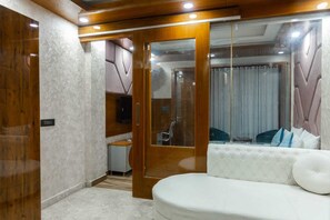 Un dormitorio moderno con un sofá blanco, un armario de madera, un televisor y un baño visible a través de una puerta de cristal.