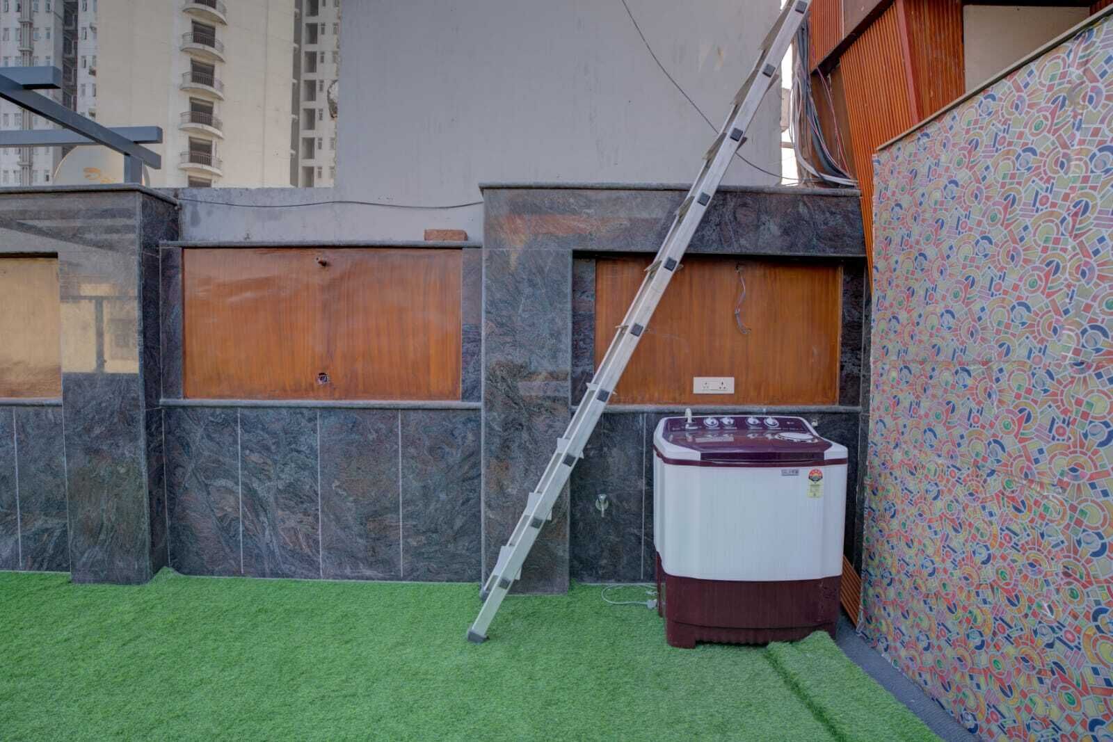 Un pequeño espacio exterior con césped artificial, una lavadora y una escalera apoyada en una pared.