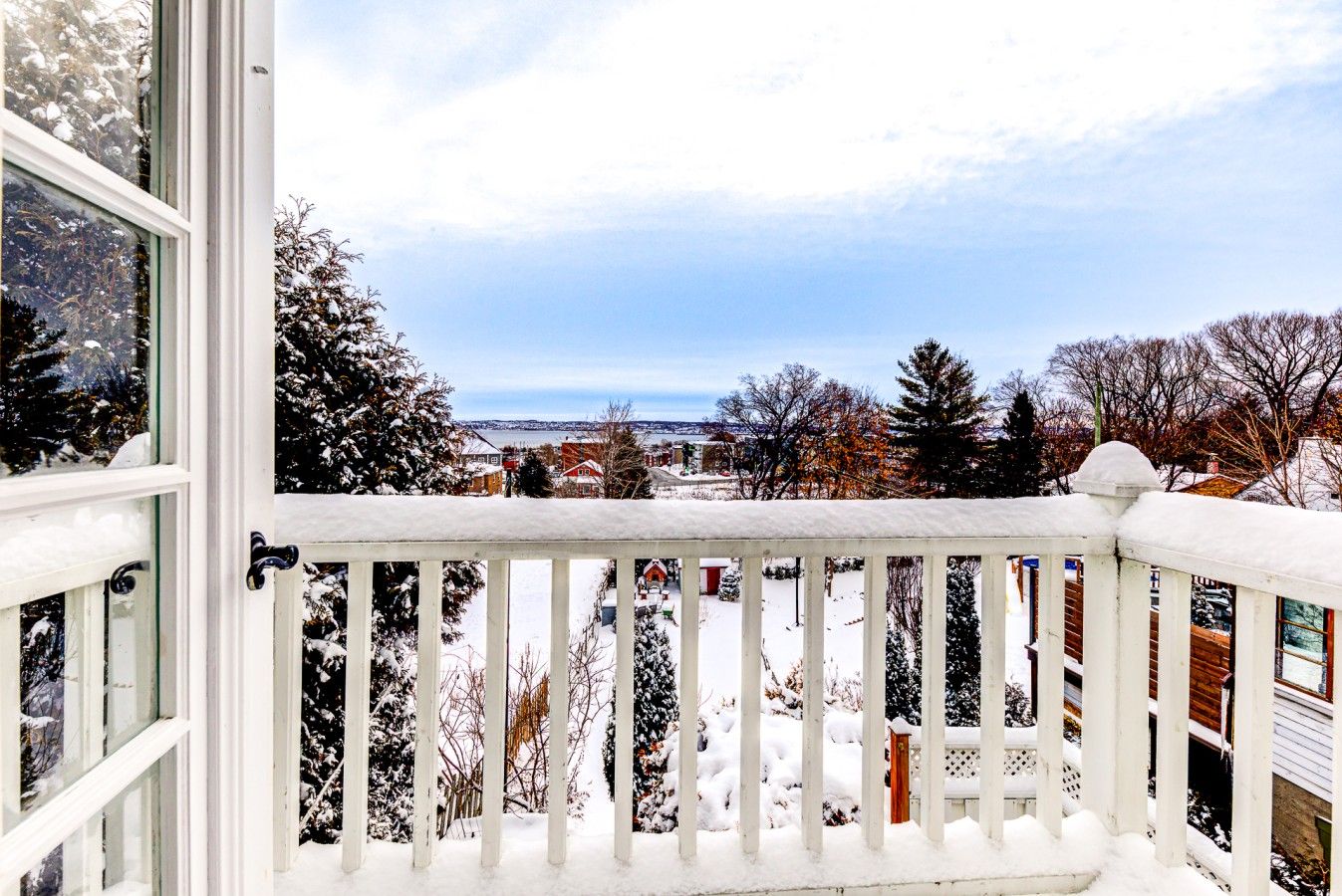 A snow-covered balcony with a view of a residential area and a body of water in the distance.